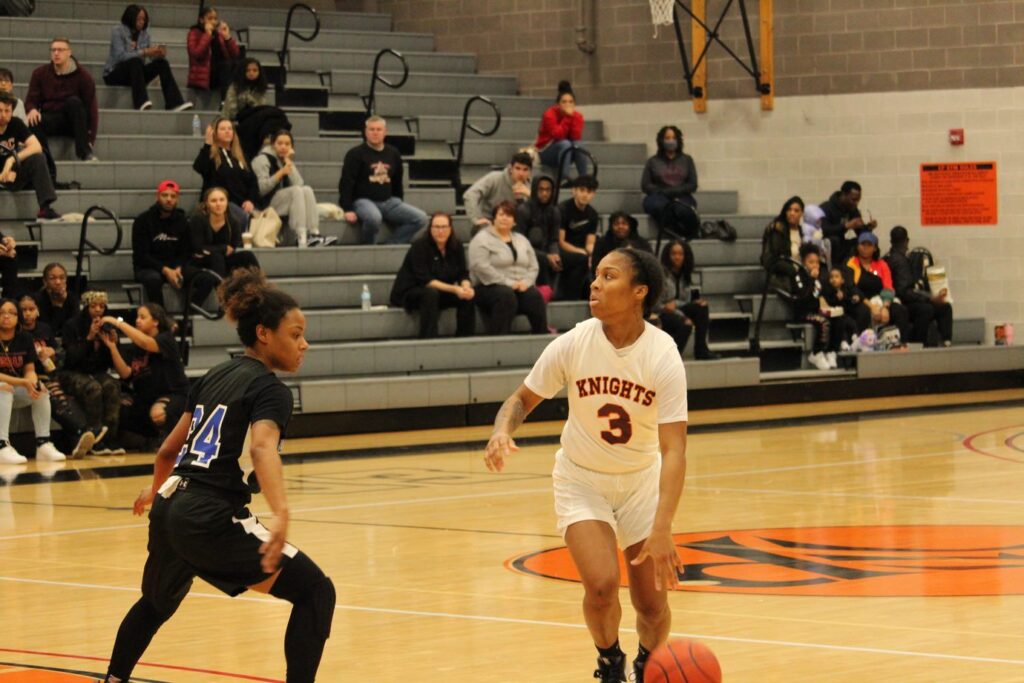 Woman basketball player, Tahniyaah Jackson, No. 3, in red-on-white uniform for the Central Penn Lady Knights dribbling the ball past half court against a defender in a black uniform, with the the blue number number 24. 