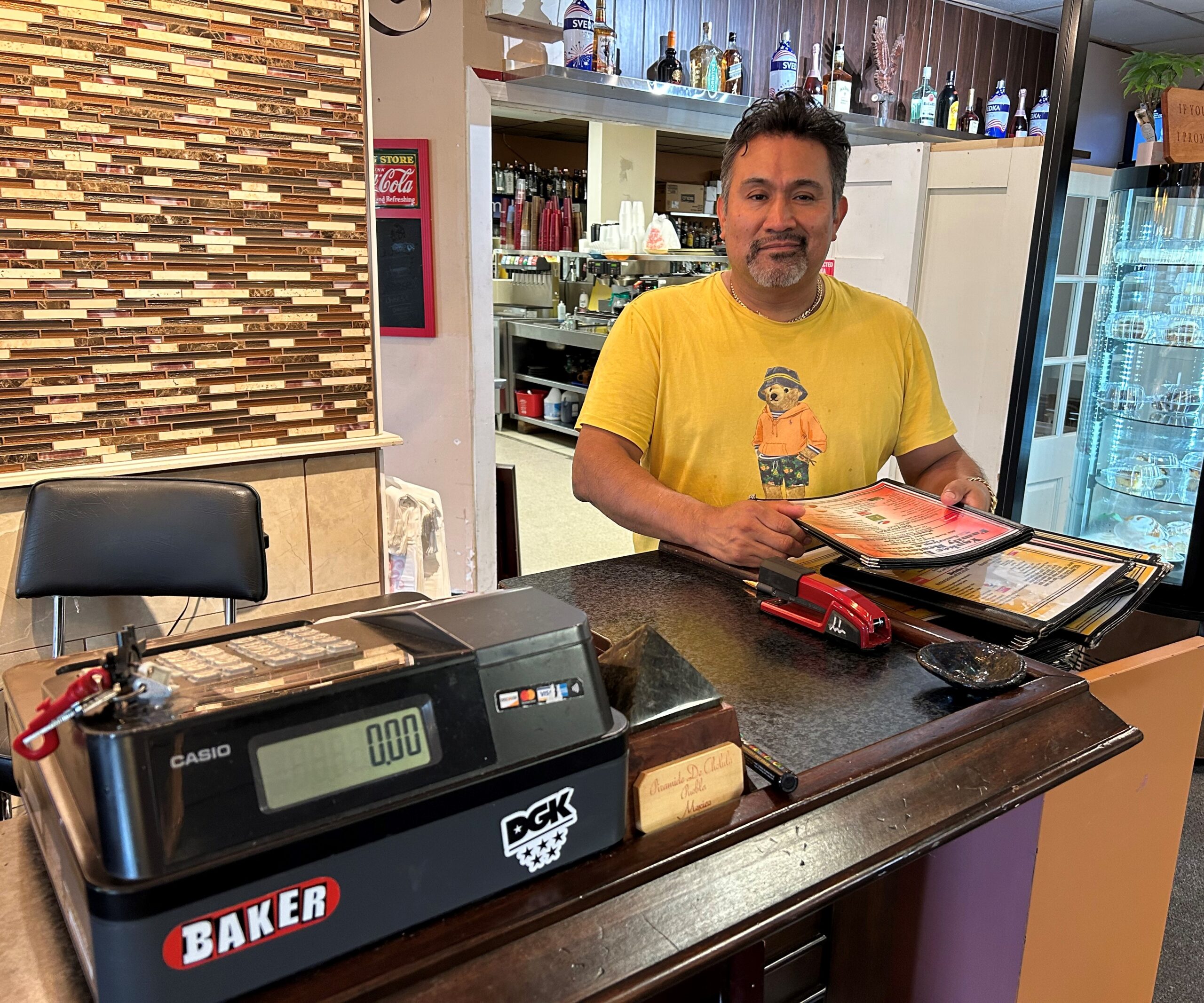 Man in yellow shirt behind a restaurant cash register counter and holding a menu