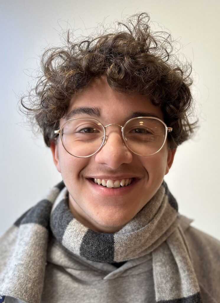 Smiling young man with curly dark hair and wire glasses, wearing a black and gray striped scarf.