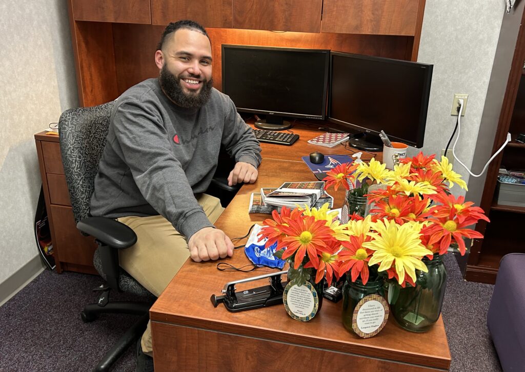 Smiling, bearded man in gray sweatshirt at a work desk with hutch.