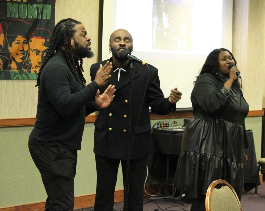 Two men and a woman dressed in black singing at the luncheon.