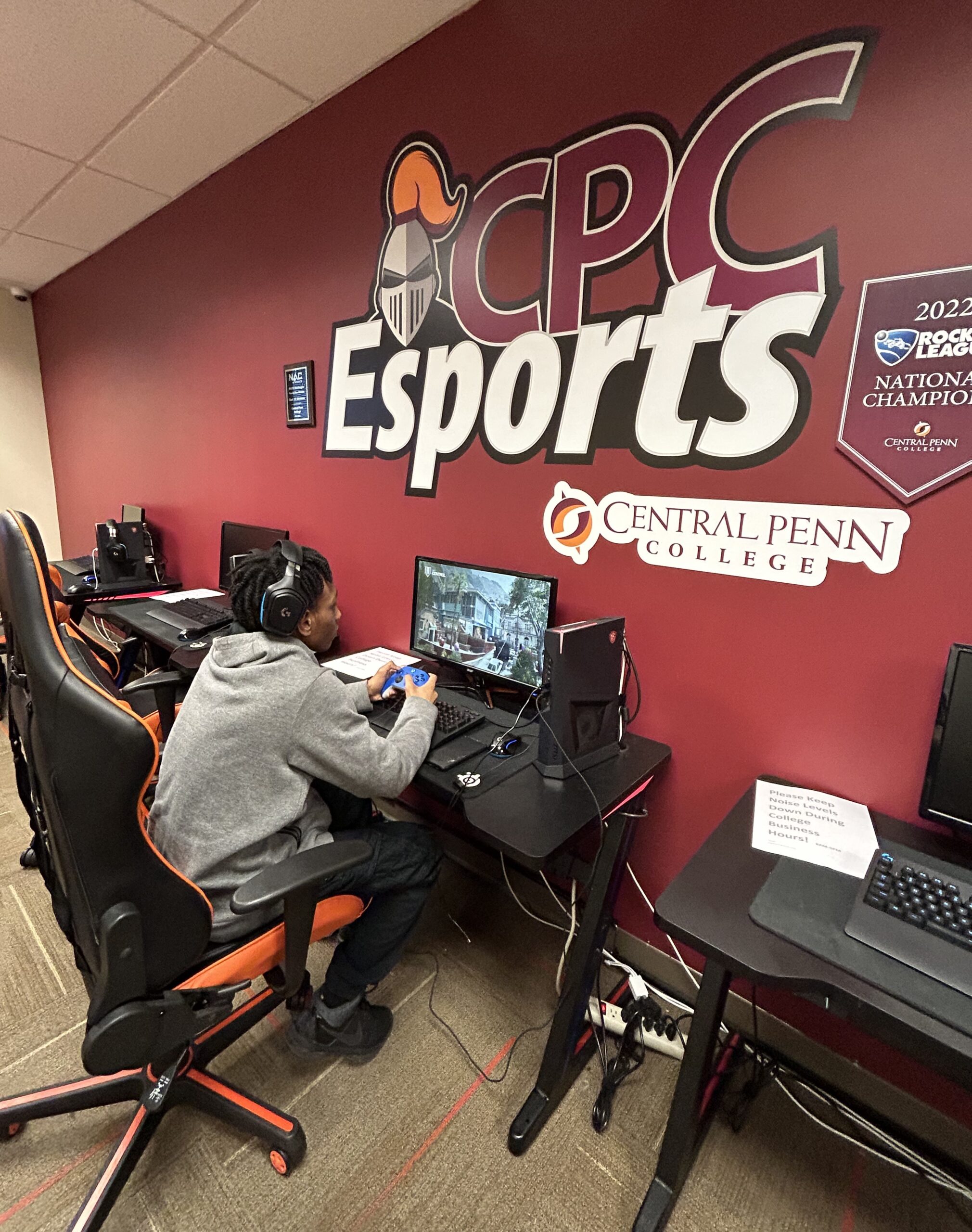 Young man in a gray sweatshirt seated in a gaming chair playing Call of Duty vanguard on a computer monitor. In front of him is a reddish-orange wall with the Central Penn knight logo (silver helmet with facemask and an orange plume on top of the helmet, and CPC Esports -- with the CPC in reddish-orange outlined in white and the Esports in bright white below the CPC, against a black background