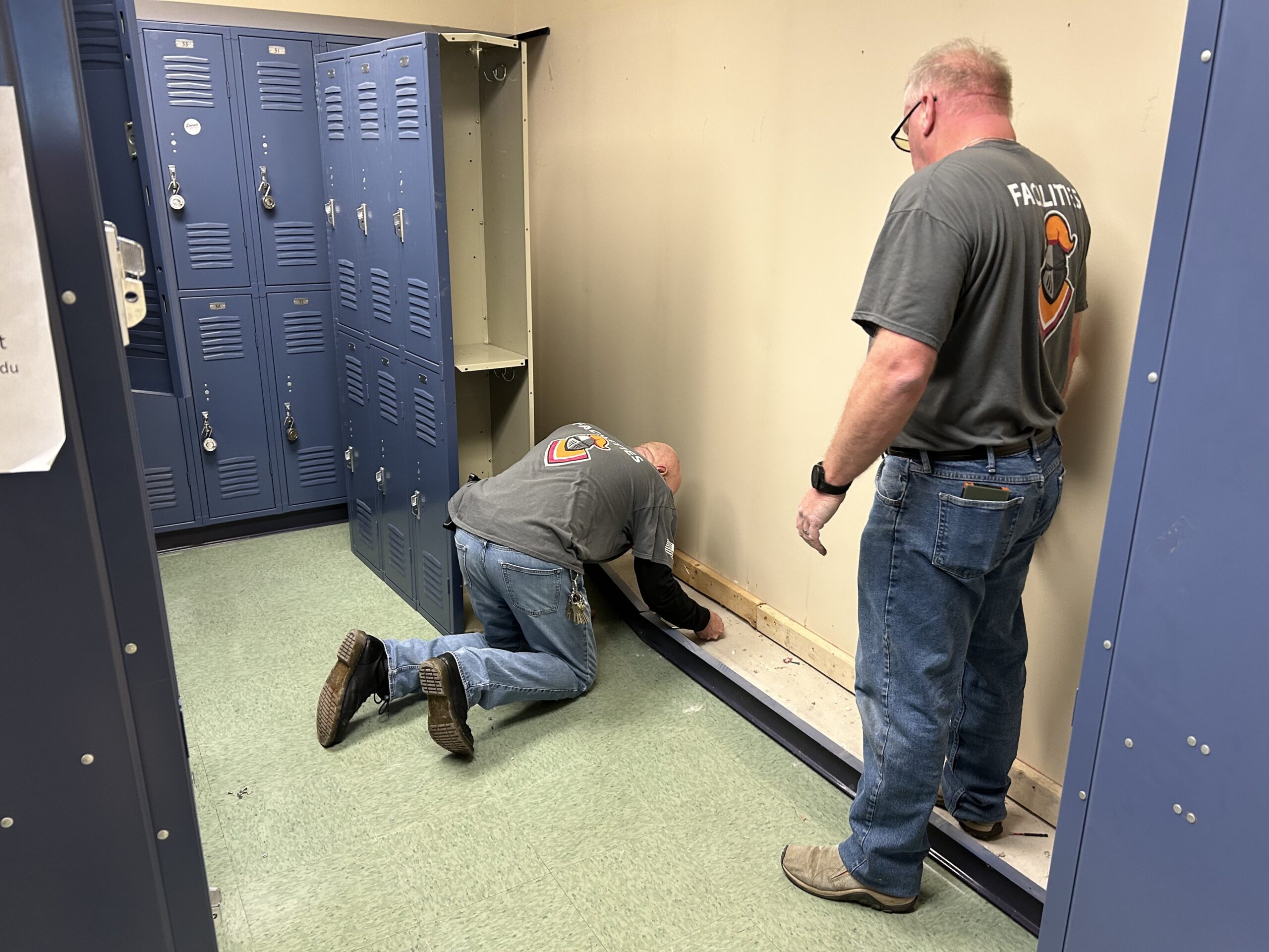 Two men in gray T-shirts and blue jeans working in a room removing double-stacked blue steel lockers.