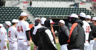A group of young-men baseball baseball players in white uniform pants and shirts with maroon numbers edged in orange. Some are wearing black jackets with orange or white flashing on the outside of the sleeves, from neck to wrist The are standing in a semicircle around a man in white pants and a black jacket with gray flashing on the sleeves and a white cap with orange visor.