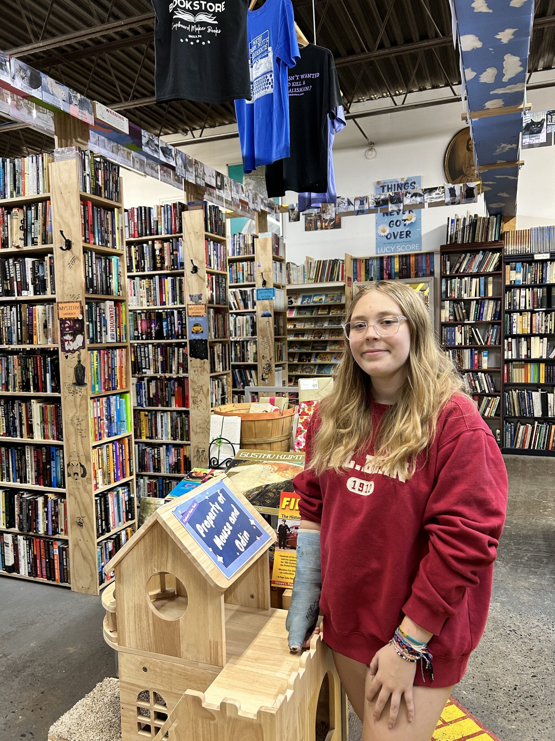Woman with long blond hair and wearing a red sweatshirt in a well stocked bookstore, and standing beside a mid-sized cat habitat with a sign on a roof side that says "Property of Mouse and Odin," two of the cats who live permanently in the Cupboard Maker Books store in Enola, Pennsylvania.