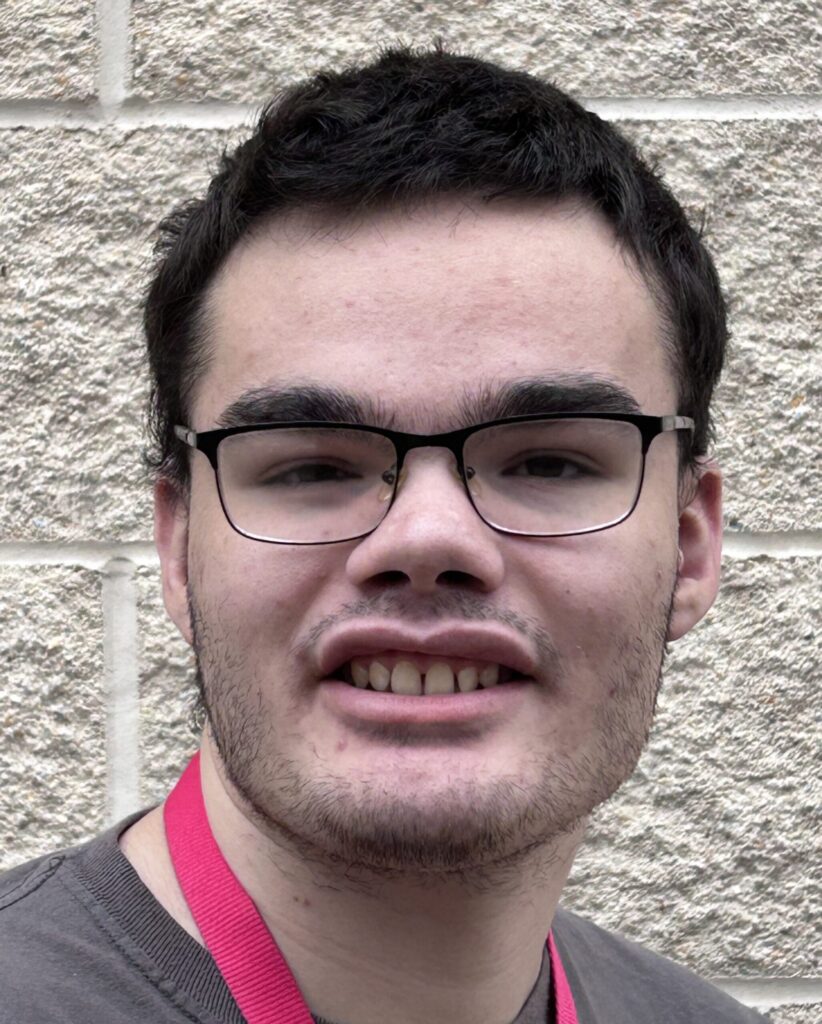 Young man smiling, with short dar, hair and dark-framed eyeglasses. Elijah Huhn