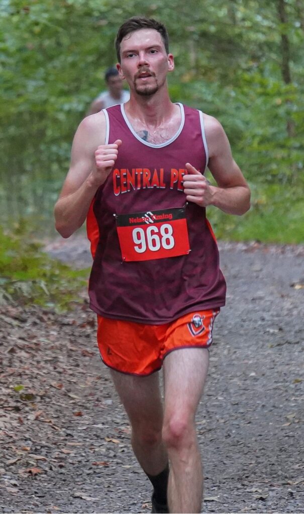 Tall thin man with short dark hair and a mustache and chin beard running on a dirt path in a wooded area. He is wearing a maroon tank top jersey with white outline on the neck and shoulder areas. Central Penn is written in thick letters across the chest of the jersey, and a large rectangular sign with 968 is pinned below the Central Penn. He's also wearing orange shorts, with a head-and-chest logo of a knight with a helnmet, on the left front thigh. Bret Savelesky running at the Penn State Brandywide Invitational cross-country meet on Oct. 5, 2024.