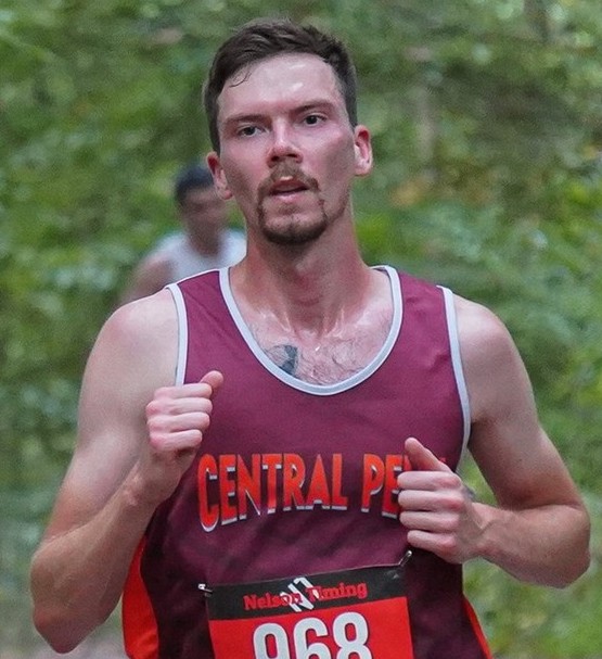 Tall thin man with short dark hair and a mustache and chin beard running on a dirt path in a wooded area. He is wearing a maroon tank top jersey with white outline on the neck and shoulder areas. Central Penn is written in thick letters across the chest of the jersey. Bret Savelesky running at the Penn State Brandywide Invitational cross-country meet on Oct. 5, 2024.