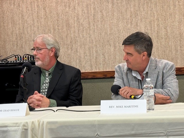 Two men sitting at a long table with a white tablecloth on it. The man on the right is Mike Martine, senior pastor at Trinity Luthern Church, Mount Joy. He has short dark hair and is wearing a light-colored open-top dress shirt and a gray-and-white tight-patterned sport coat over it. He's hold a microphone with a black foam wind cover on it. On the left is Chad Hogue, senior pastor at New Hope Church (Brethren in Christ), Harrisburg. He has short white hair and a sparse white beard and mustache, along with dark-framed eyeglasses, and is wearing a green dress shirt and green tie with while spots, and a black suit coat.