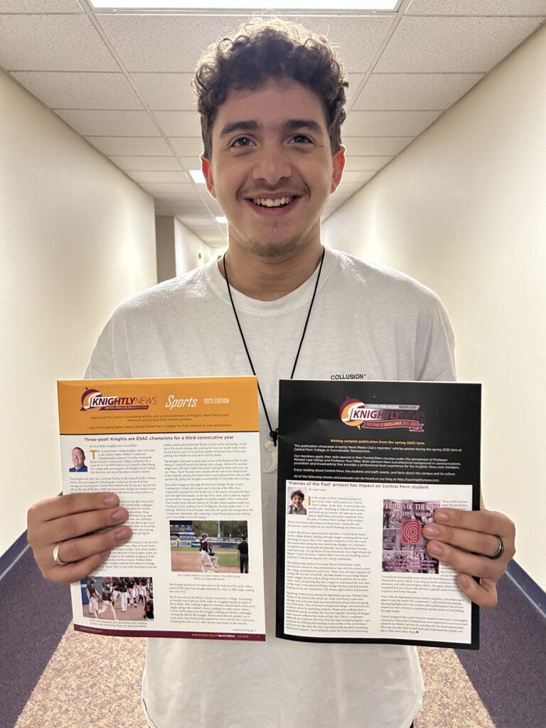 Young smiling man with short curley hair holding two Knightly News printed commemnorative editions, one in each hand, and displaying them for onlookers, Noah Lopez