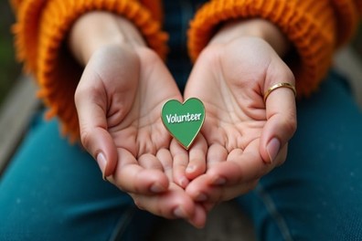 Hands cupping a gold-rimmed green heart-shaped pin that has "Volunteer" written in the middle in white.