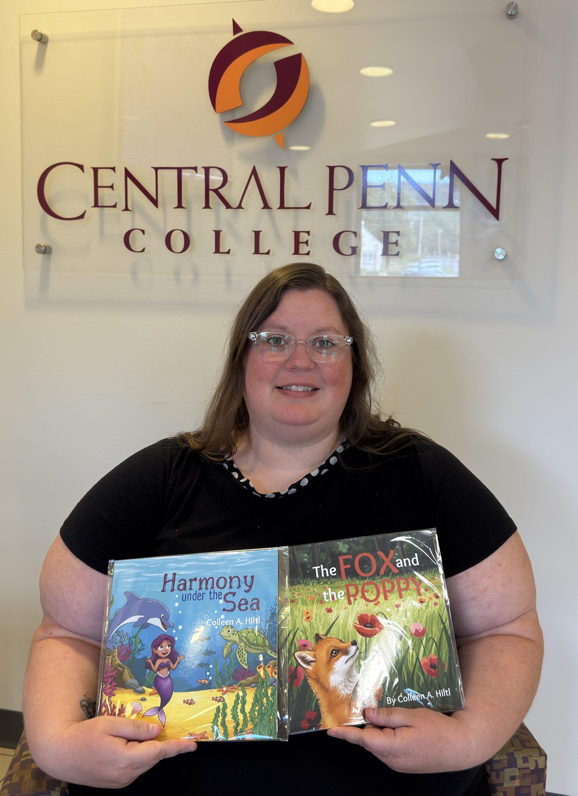 Smiling woman with long dark-blond hair and wearing clear-framed eyeglasses and a black dress, with a white polka-dot neck line, sitting on a bench in the lobby of Central Penn College's Advanced Education Technology Center building, holding on her lap her two children's books, "The Fox and the Poppy" and "Harmony Under the Sea."