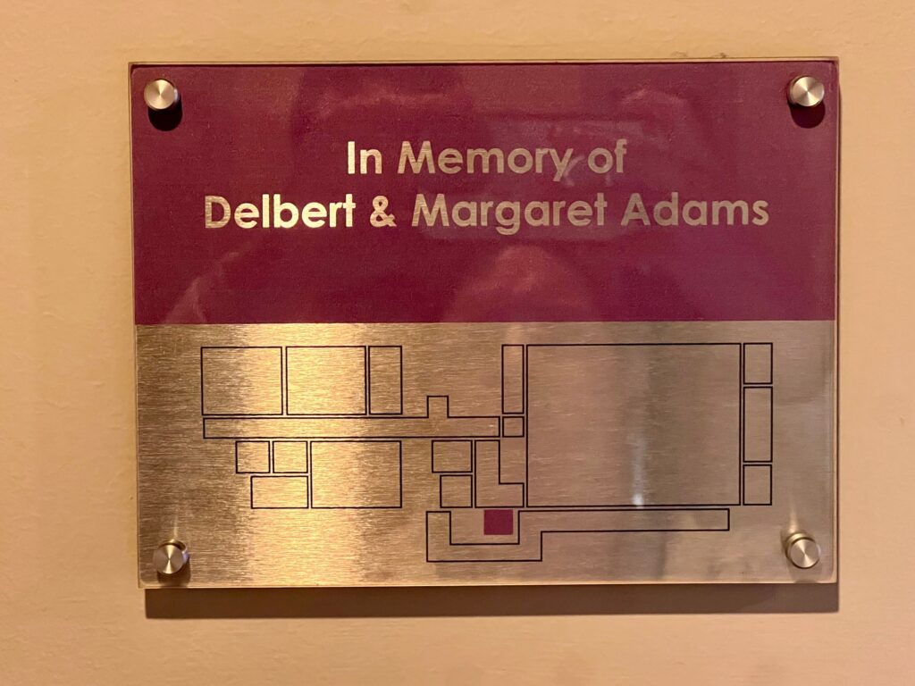 A glossy thin bronze or other type small metal square plaque, gold onh the bottom and red on top (half and half), affixed to a tan wall with four gold-stopped bolts. Inscribed in the red field on top is In Memory of Delbert and Margaret Adams. Etched into the gold field on the lower half is a line-drawing outline of the floor plan of the Central Penn College Underground, which contains a black-box theater, some small storage and other rooms, including a kitchenette, a large student lounge, two restrooms, a small exercise room, a small dance-practice studio and a few offices.  