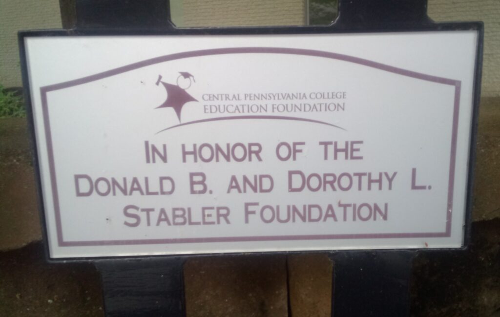 Small rectangular plaque on a bench. White with black border and red imprint on front, in a bed headboard design in red, with Central Pennsylvania College Education Foundation In Honor of Donald B. and Dorothy L. Stabler Foundation. It's afixed to a slatted bench.