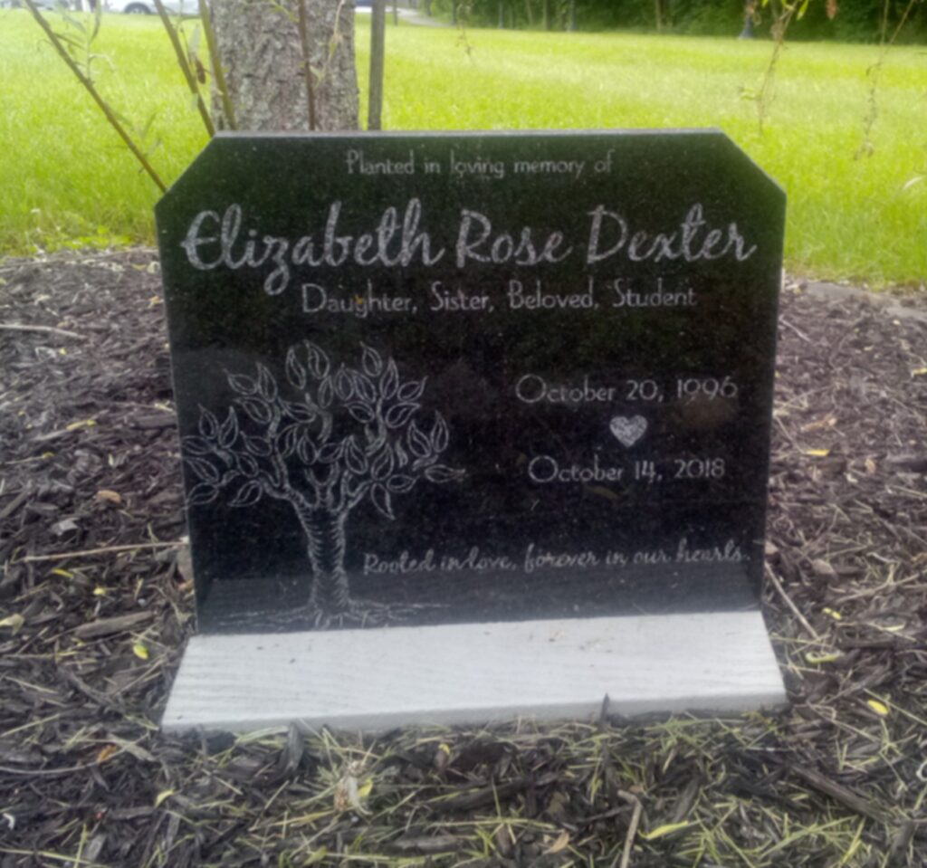 A black stone memorial marker in the shape of a squarish tombstone. It has a whuite flat horizontal foundation sticking out about a foot from the front. It is in front of a tree, the lower part of the truck visible in this phtoograph, behind the upper left edge of the memorial stone. On the stone is etched in script at the top, in light gray, Planted in loving memory of. Below it, in large light-gray script in cursive, is Elizabeth Rose Dexter. Below that, also in light-gray cursive, is Daughter, Sister, Beloved, Student. Below that to the right (looking at the photograph) is October 20, 1996, with a light-gray solid heart carved in the middle immediately below the 20, and below that October 14, 2018. On the left side of the stone (looking at it on the blog page) is an etching in light gray of a small, stout-trunked tree with pl;entiful branches with large single leaves. Next to the tree, on the right, at the bottom of the marker, in light-gray cursive script, is Rooted in love, forever in our hearts.