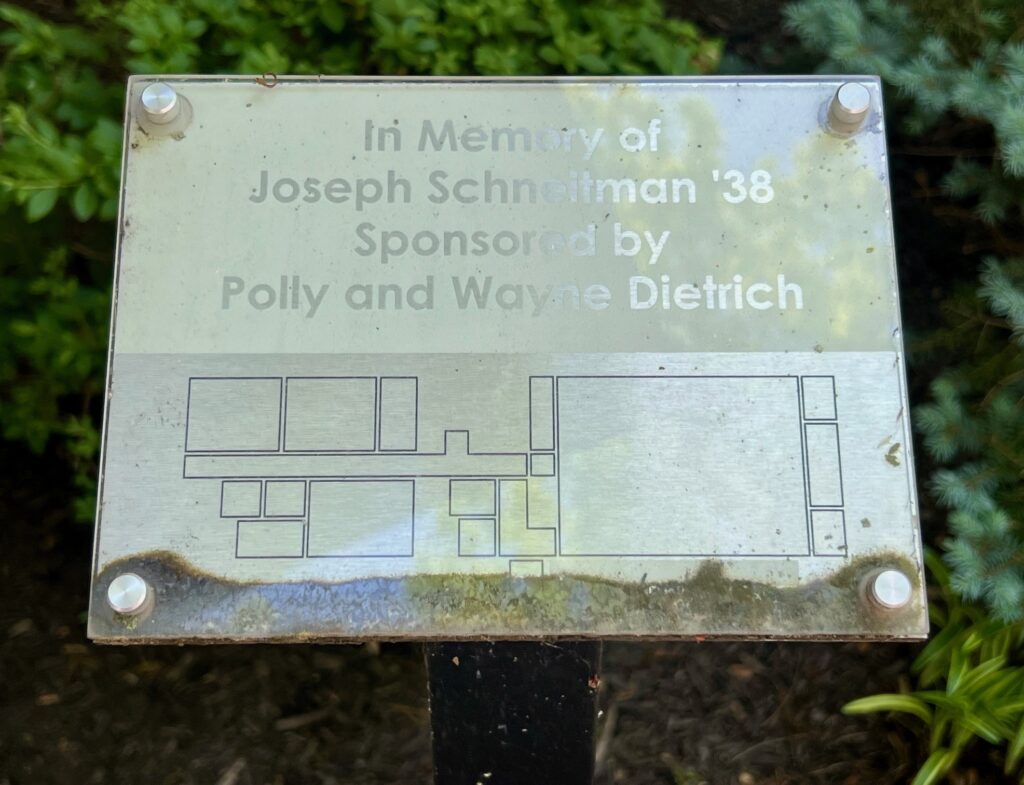 A small square plaque, material unknown. It is affixed to the top of a small short sticklike post in a garden bed on the Central Penn College Campus. The top half is a whitish-gold opaque background imprinted with In Memory of Joseph Schneitman 38 Sponsored by Polly and Wayne Dietrich. On the bottom half, which is white and opaque, is inscribed a line-drawing outline of the floor plan of The Underground at Central Penn, below the West Wing of Milano Hall. The Underground contains a black-box theater, some offices and other small rooms, two restrooms, a large student lounge, an exercise center and a small practice dance studio.