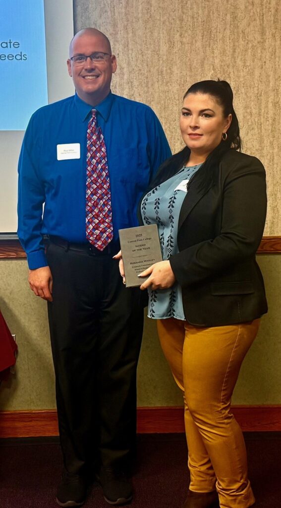 Man with shaved head and wearing eyeglasses and a dark-blue dress shirt with a red-and-whie patterned tie standing next to a woman with pinned-back long black hair and wearing a black blazer and gray blouse with a black vertical leaf patter. The woman is holding a plaque recognizing her as Intern of the Year, presented at the Central Penn College Business Partner of the Year Breakfast on Sept. 11. The man is Prof. Paul Miller, academic coordinator of communications internships, and the woman is senior corporate-communications student Rosanna Wakley.