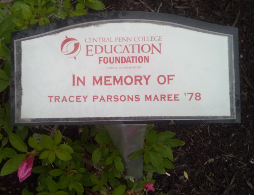 Small memorial plaque that is horizontally rectangular, with the upper border curved gently up in the middle. It has a white background and a gray border. In the middle, in red letters and numbers, is Central Penn College Education Foundation In Memory Of Tracey Parsons Maree '78.