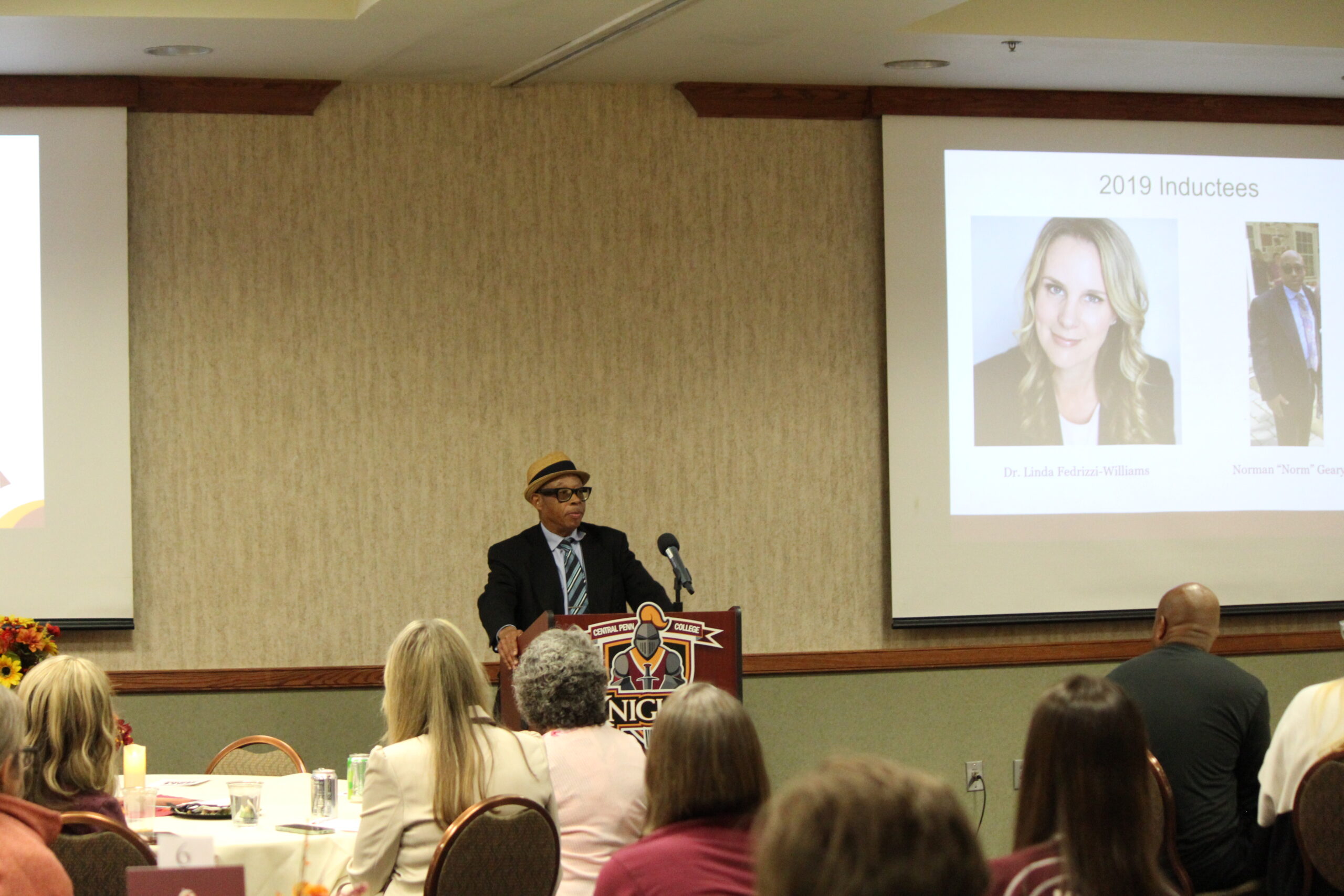 Man with hat addressing a room of people
