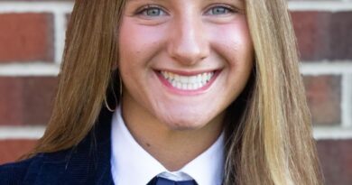 Smiling young woman with a deep-blue high school varsity-type jacket and also wearing a white dress shirt with a medium-dark blue necktie. Alana Peiffer