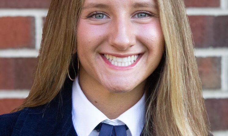 Smiling young woman with a deep-blue high school varsity-type jacket and also wearing a white dress shirt with a medium-dark blue necktie. Alana Peiffer