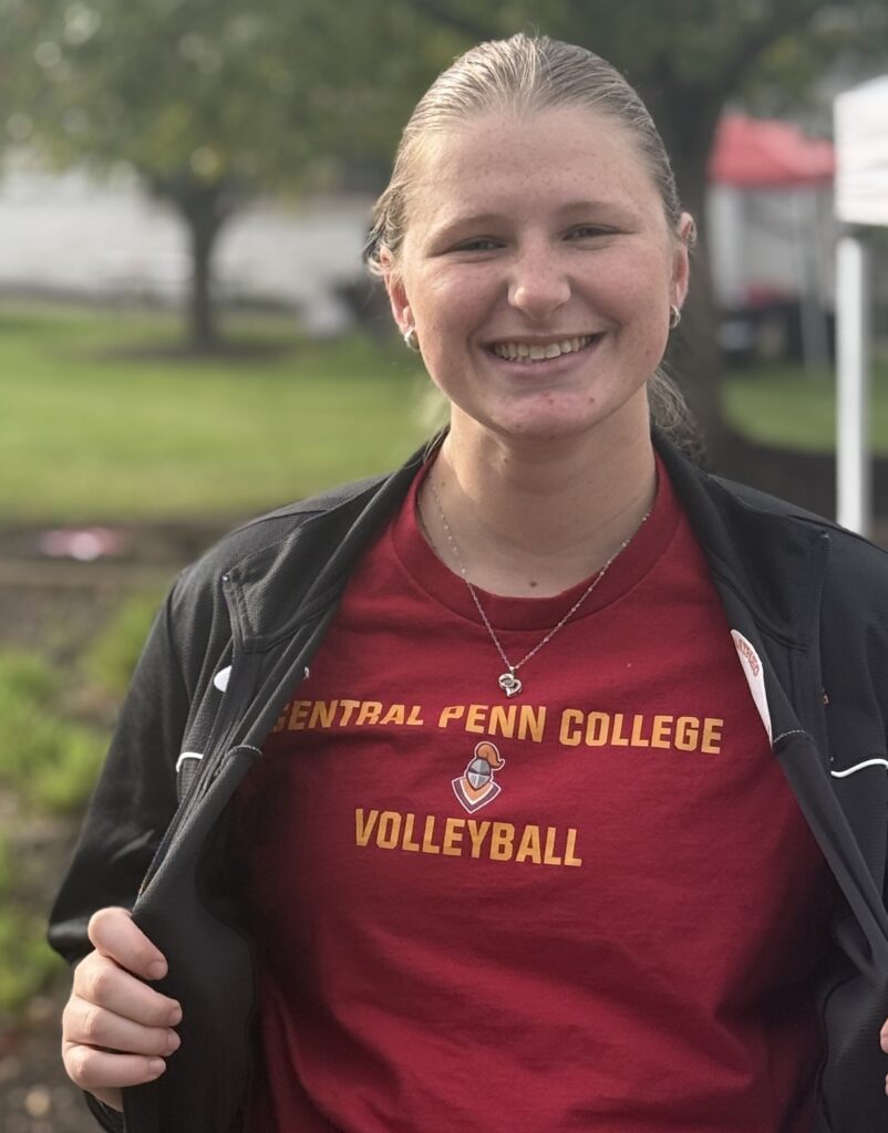 Young woman with tied-back blond hair wearing an open black windbreaker jacket and a maroon T-shirt with gold print that says Central Penn College Volleyball