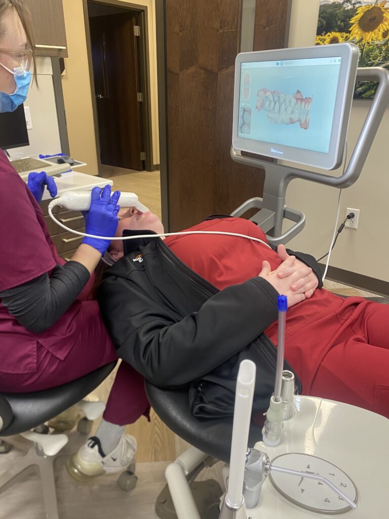 A woman student, wearing maroon medical scrubs, hair in a ponytail, uses a 3d intraoral scanner to capture images of the inside of another student's mouth as the second student lies on her back in a dental chair. An image of a ring of teath is displayed on a monitor next to the students.