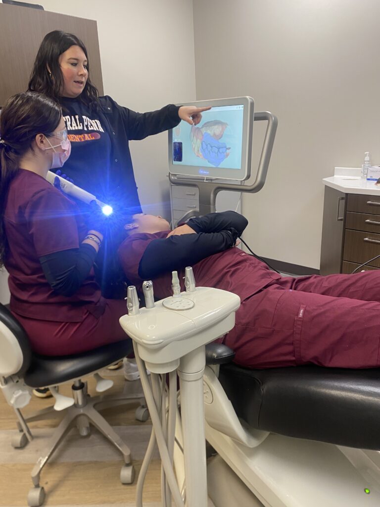Two women students in red medical scrubs. One is lying on her back in a dental chair. The other is is sitting next to her, holding an intraoral scanner that is shining a small globe of bright blue light, with a blue aurora. A woman with long dark hair is standing near them, discussing an image on a computer screen next to them, at which the students are looking. 