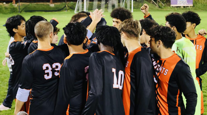 A group of young men in black and orange jerseys in a tight huddle, their hands up. Central Penn Knights men's soccer team.