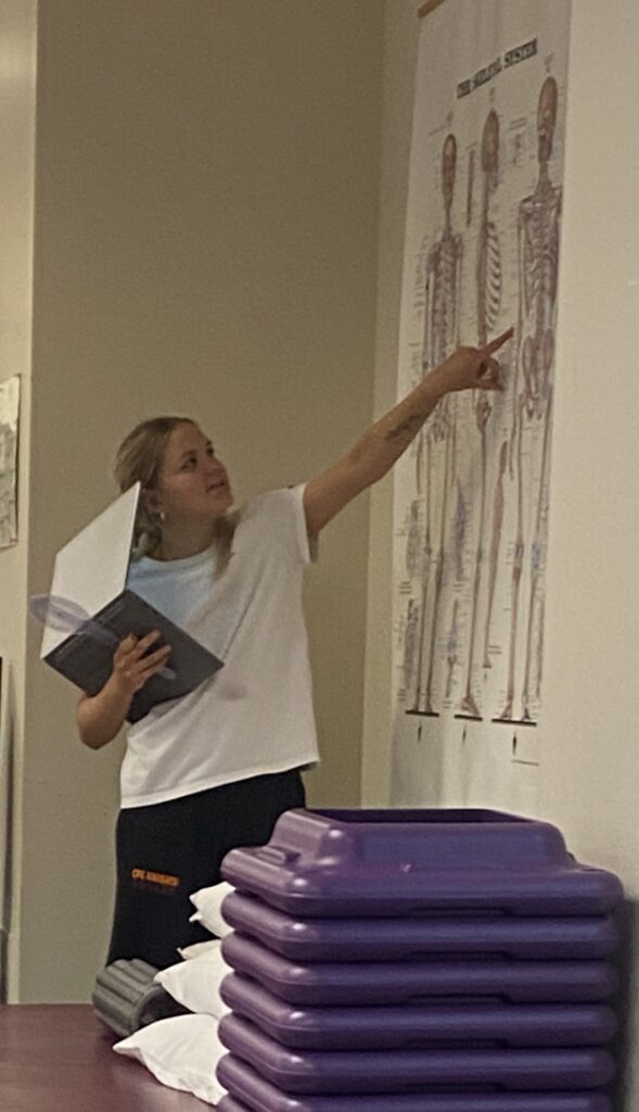 Woman with blond hair pointing to a skeletal anatomy chart on a wall in a lab.