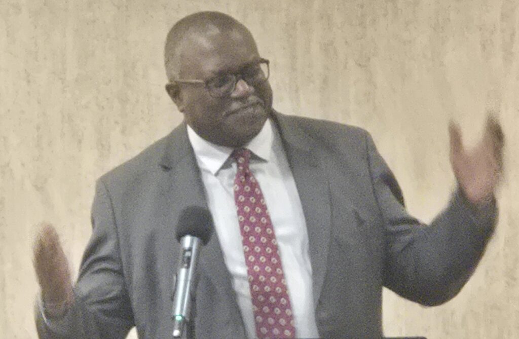 Man n gray suit, with arms apart to the sides halfway, and wearing a white dress shirt and a red tie with a white round pattern on it,