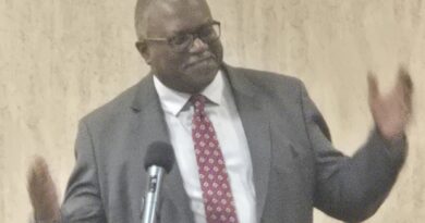 Man n gray suit, with arms apart to the sides halfway, and wearing a white dress shirt and a red tie with a white round pattern on it,