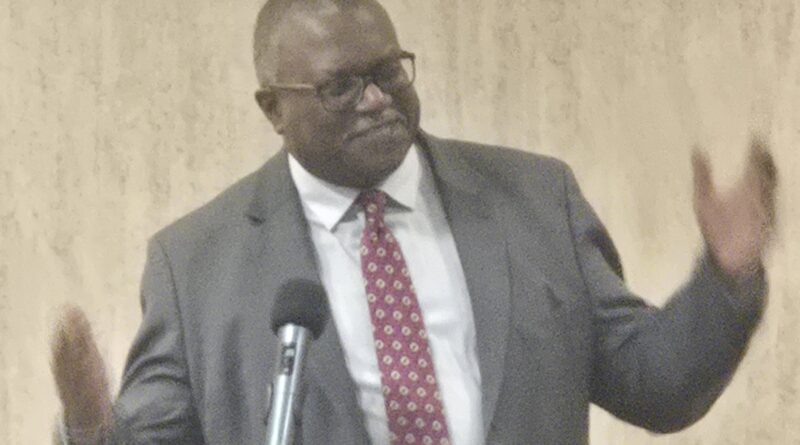 Man n gray suit, with arms apart to the sides halfway, and wearing a white dress shirt and a red tie with a white round pattern on it,