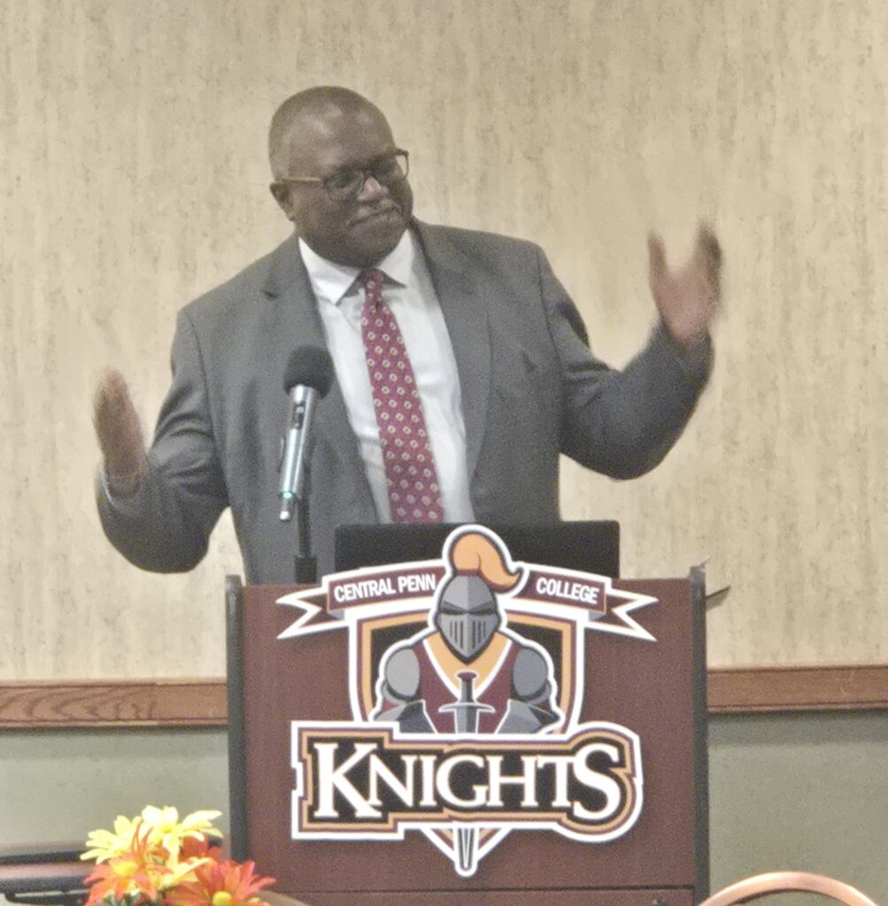 Man in a gray suit behind a podium gesturing, with arms halfway out from his sides. Joseph Robinson.