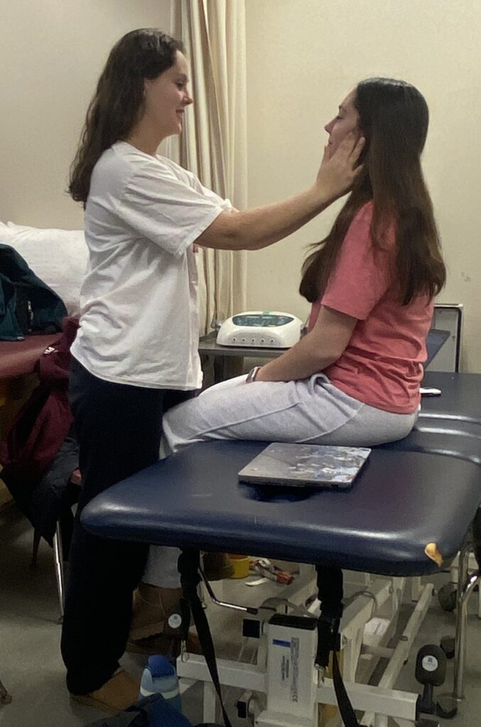 One woman standing and examing the jaw and face of another woman sitting on a table.