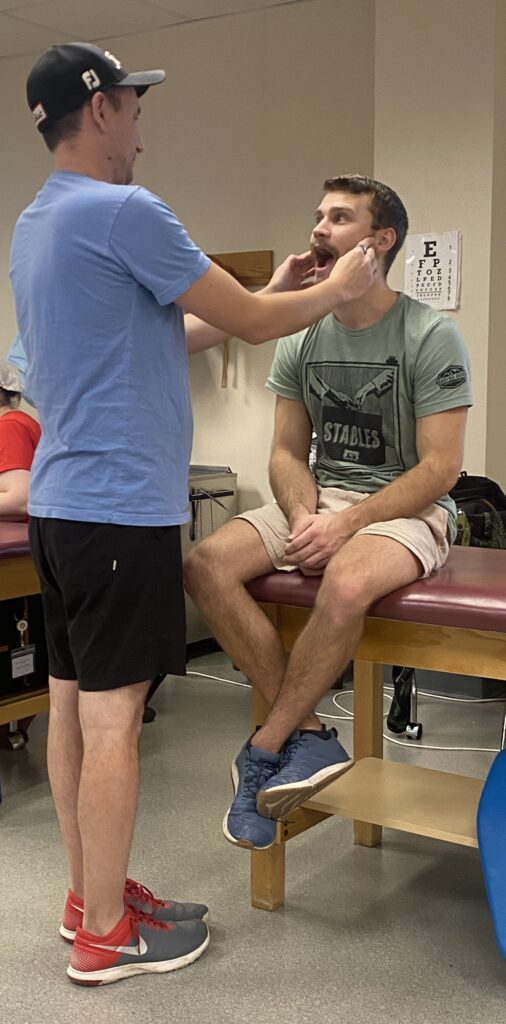 Man standing and measuring how far and easily a man seated on an examination table can open and close his mouth.