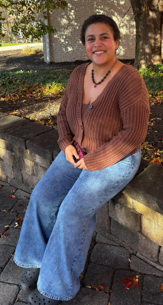 Smiling young oman with short dark hair sitting on a low stone wall on Central Penn's campus, in the quad. Jalicia Johnson.