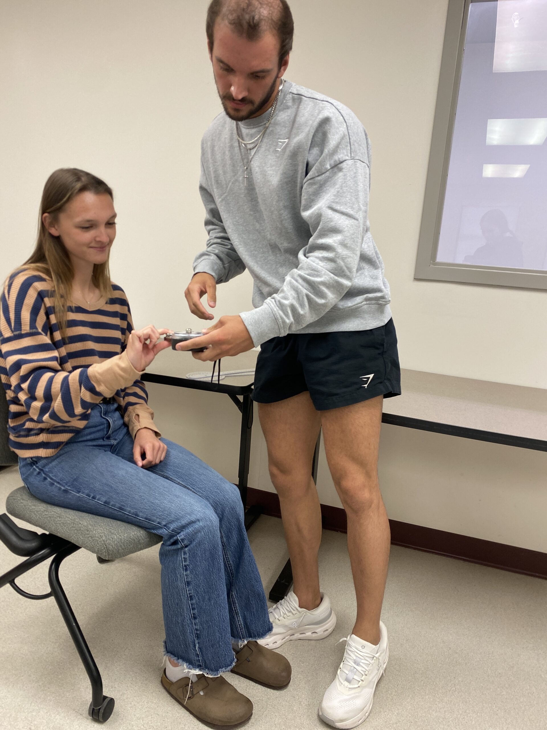 A man in a long-sleeved shirt and shorts working with a woman in a striped sweater and blue jeans, the latter who is sitting on a chair, in the Central Penn OTA lab.