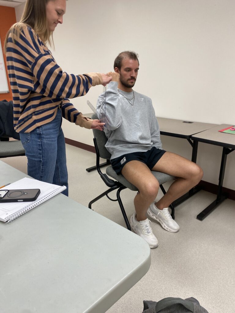 Woman in horizontal-striped sweater and blue jeans standing behind a man in a long-sleeved shirt and shorts who is sitting in a chair. She is taking measurements in the OTA lab.