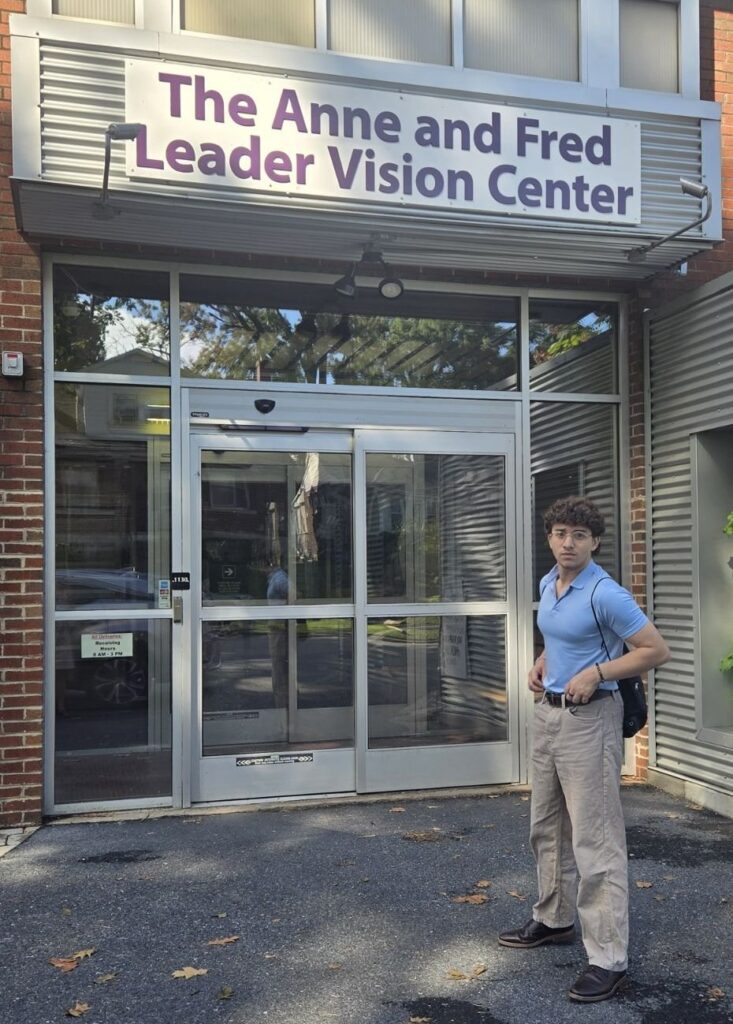 You man with mid-dark curly hair and wire-framed eyeglasses, and wearing a light-blue button-V neck business-casual polo short and tan jeans, standing in front of the Anne and Fred Leader Vision Center.