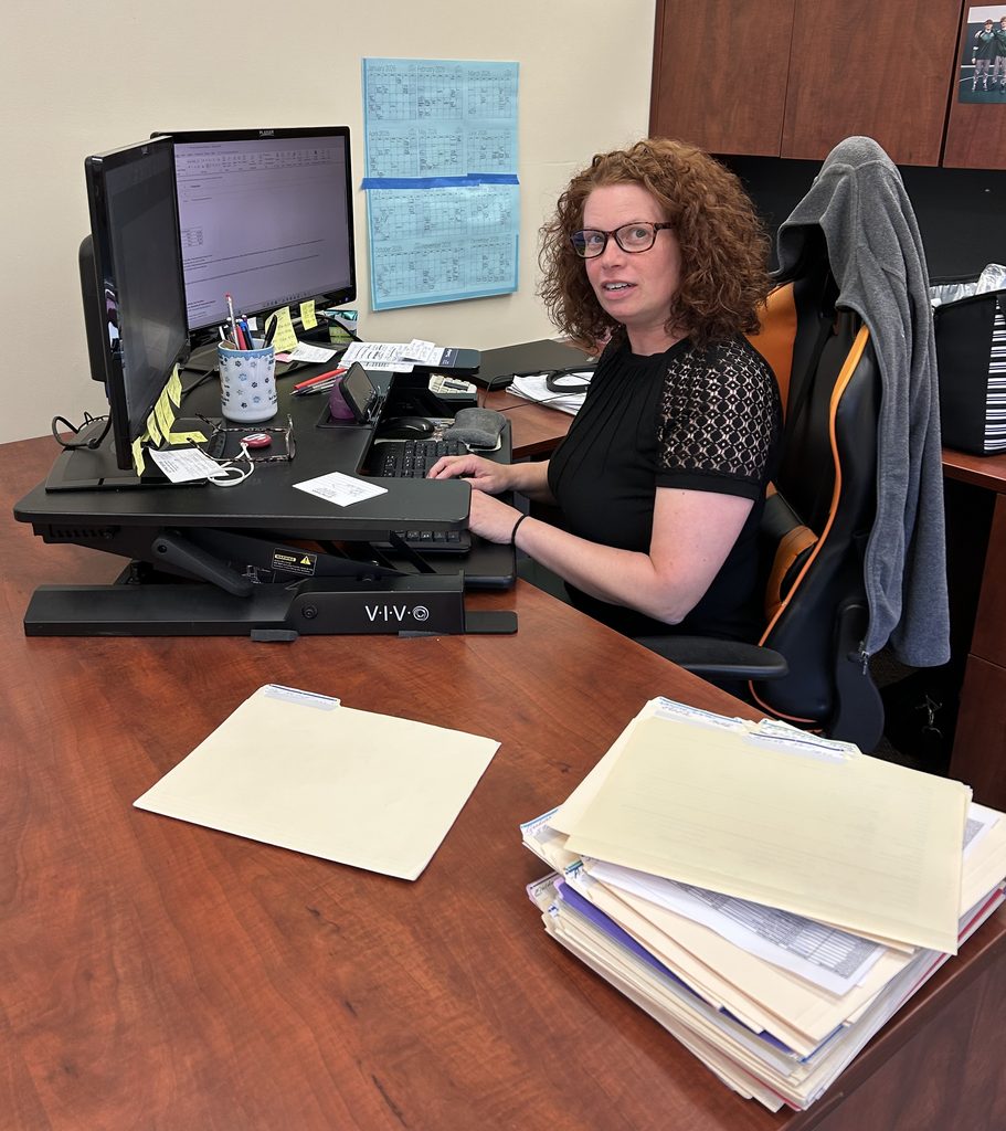 Woman with shoulder-length curly red hair who is wearing eyeglasses and a black short-sleeved top with sleeves printed with white dots and Xs at a double-monitored computer and work desk, with dull-yellow file folders on it Central Penn College Financial Aid Director Kathy Sherpard.