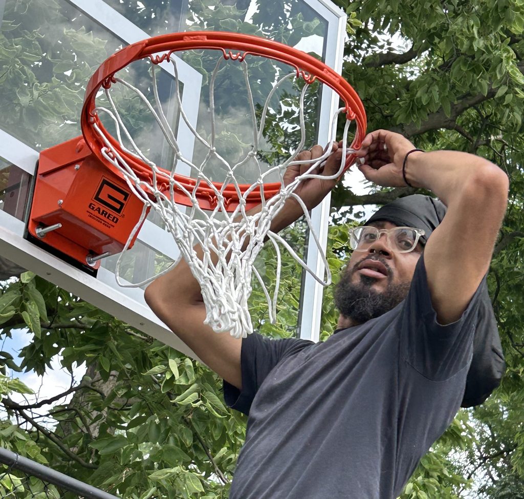 Man in black T-shirt, dark beard and eyeglasses, and wearing a black head scarf, finishing the hooking of a white net onto an orange basketball hoop affixed to a clear tempered-glass backboard.