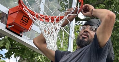 Man in black T-shirt, dark beard and eyeglasses, and wearing a black head scarf, finishing the hooking of a white net onto an orange basketball hoop affixed to a clear tempered-glass backboard.