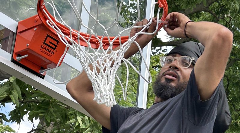 Man in black T-shirt, dark beard and eyeglasses, and wearing a black head scarf, finishing the hooking of a white net onto an orange basketball hoop affixed to a clear tempered-glass backboard.