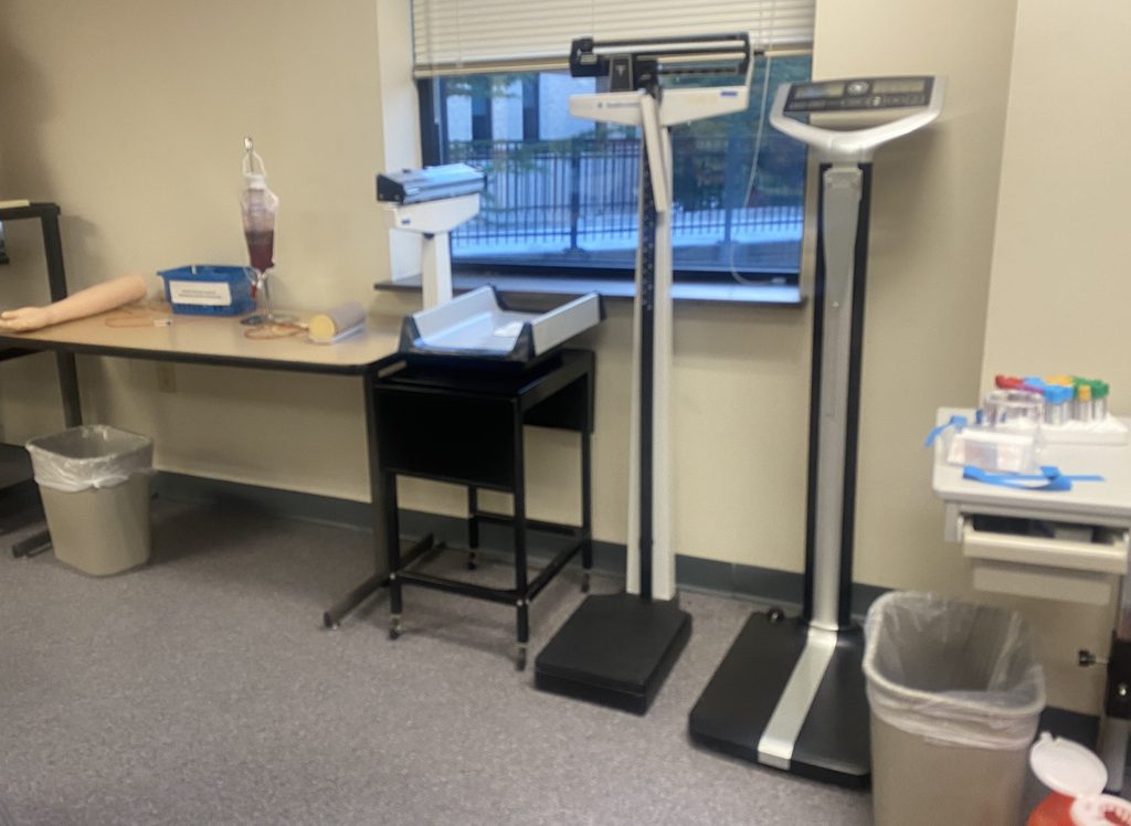 An artificial human arm on which medical-assisting students can practicing drawing blood  rests on the end of the table, at left, in the Central Penn MA lab . The table also has a blood bag to hang from an IV stand and a scale for weighing infants. Standing to the right of the table are  two medical scales--one manually operated and has sliding counterweights at the top and one digital scale. To the right of the scales is a small table holding medical-office supplies. Photo by Jules Quare